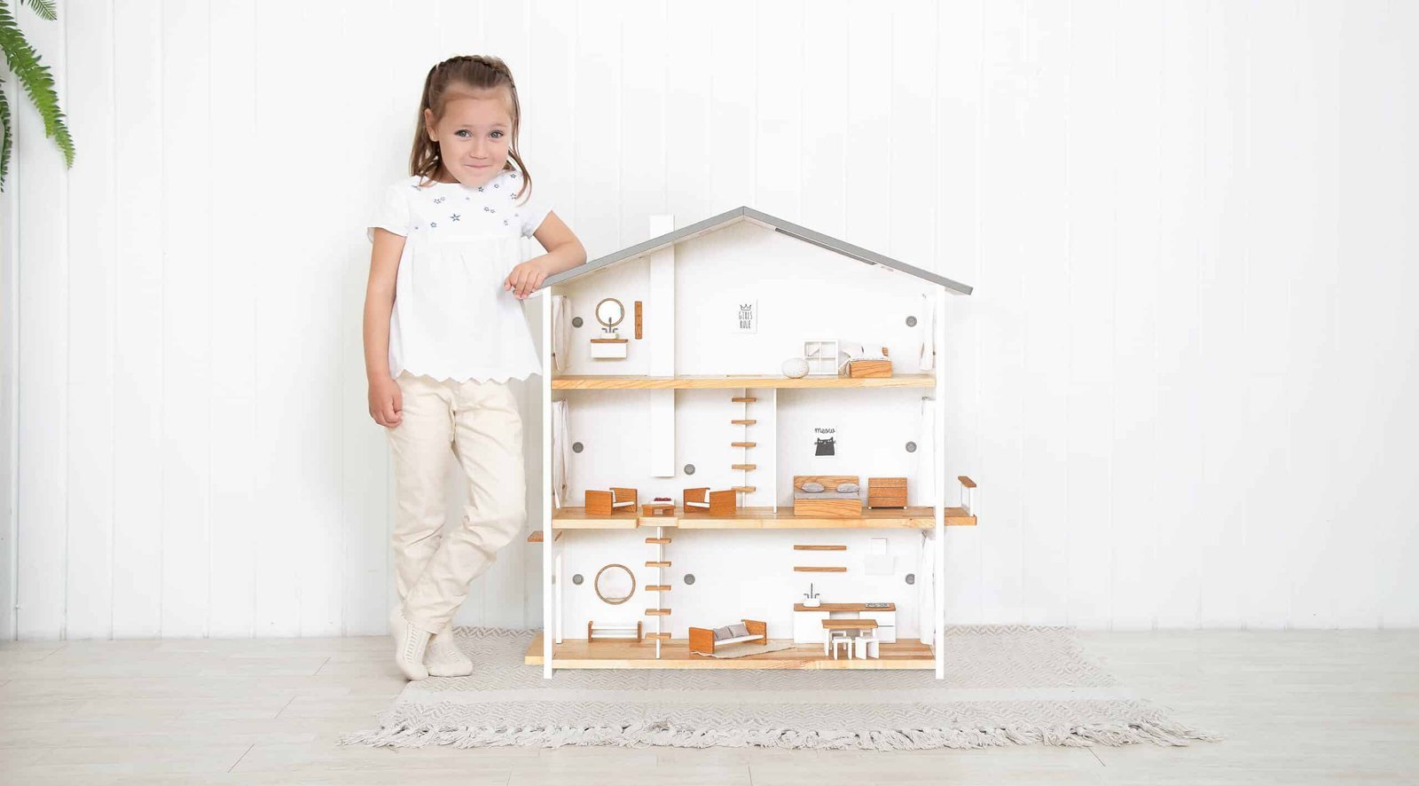 Happy 6-year-old girl leaning on a large Katin Domik wooden dollhouse in a minimalist Scandinavian interior