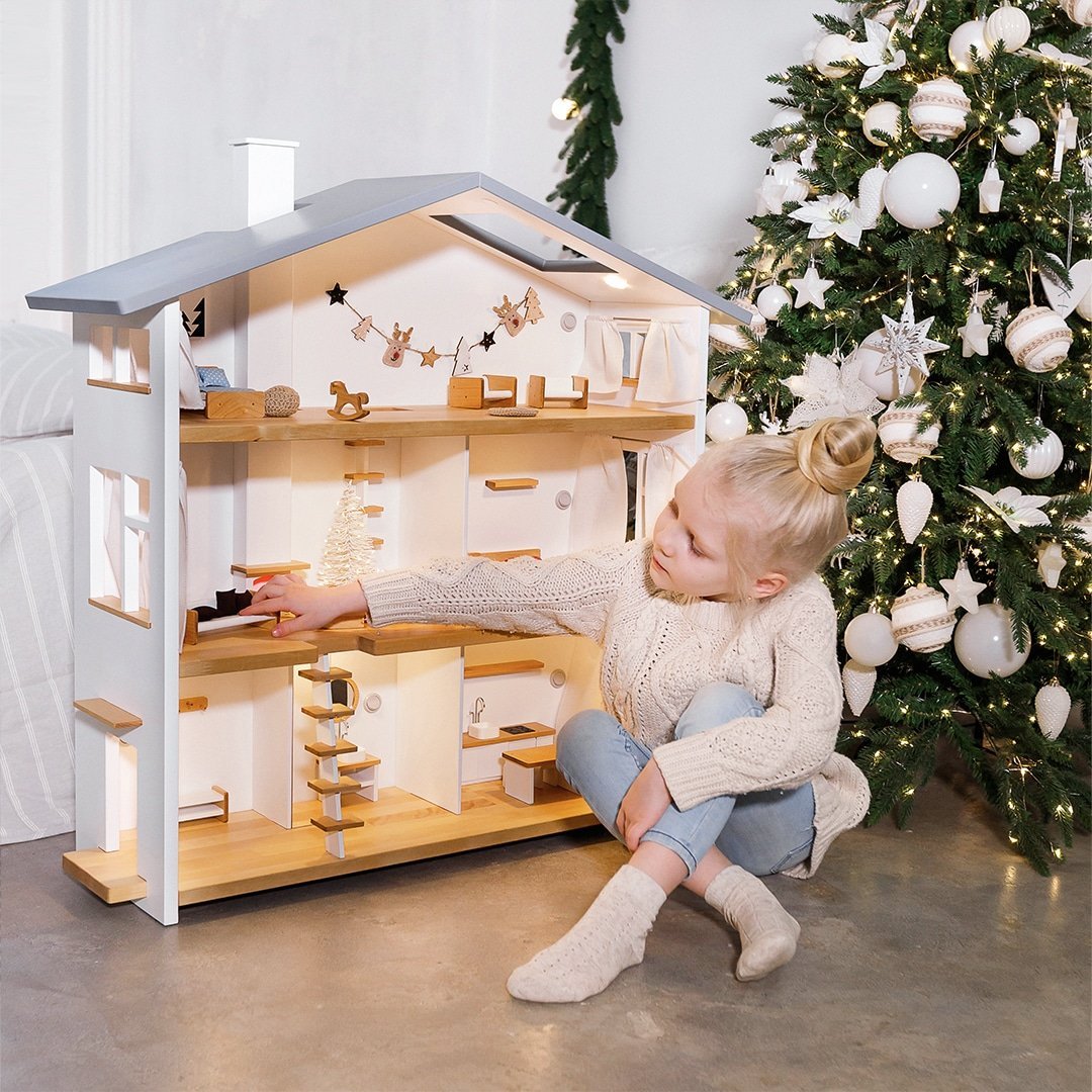 Little girl playing with a lit wooden doll house with lights next to a Scandinavian-style Christmas tree