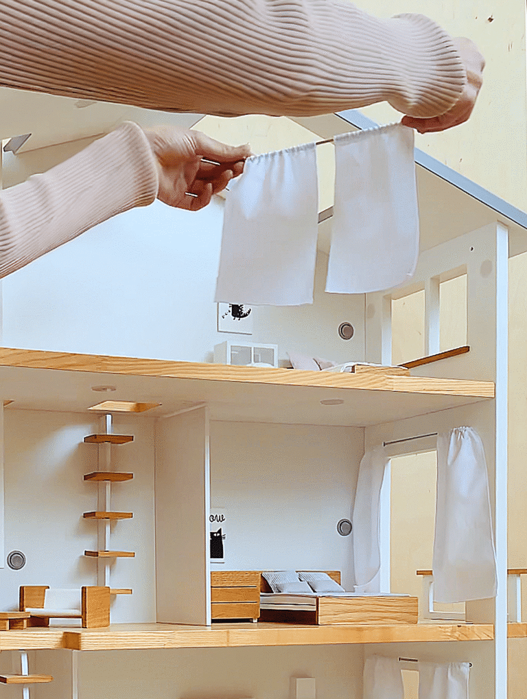 Two hands holding a removable curtain rod with fabric curtains in front of a furnished wooden dollhouse