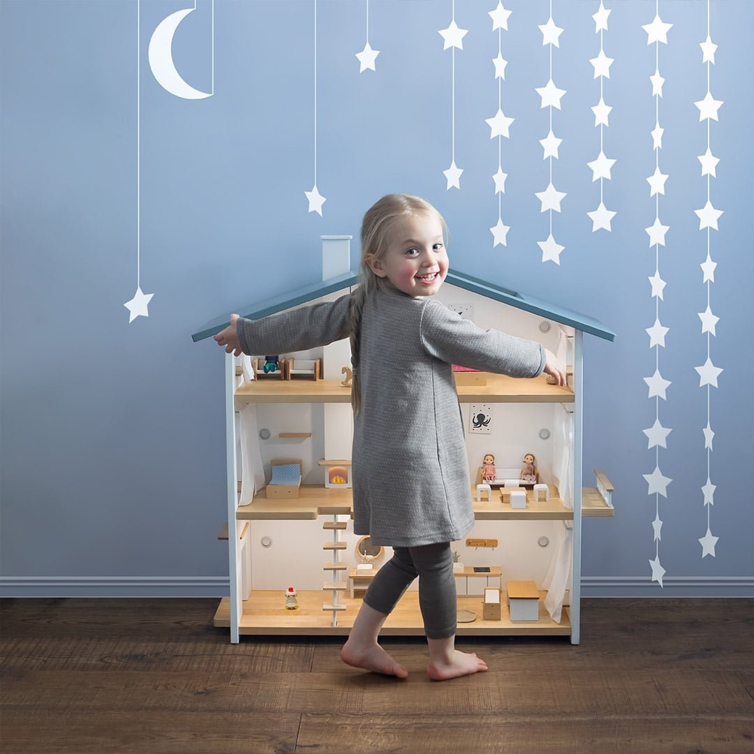 Girl hugging a wooden dollhouse with lights, surrounded by paper star and moon garlands – Christmas gift